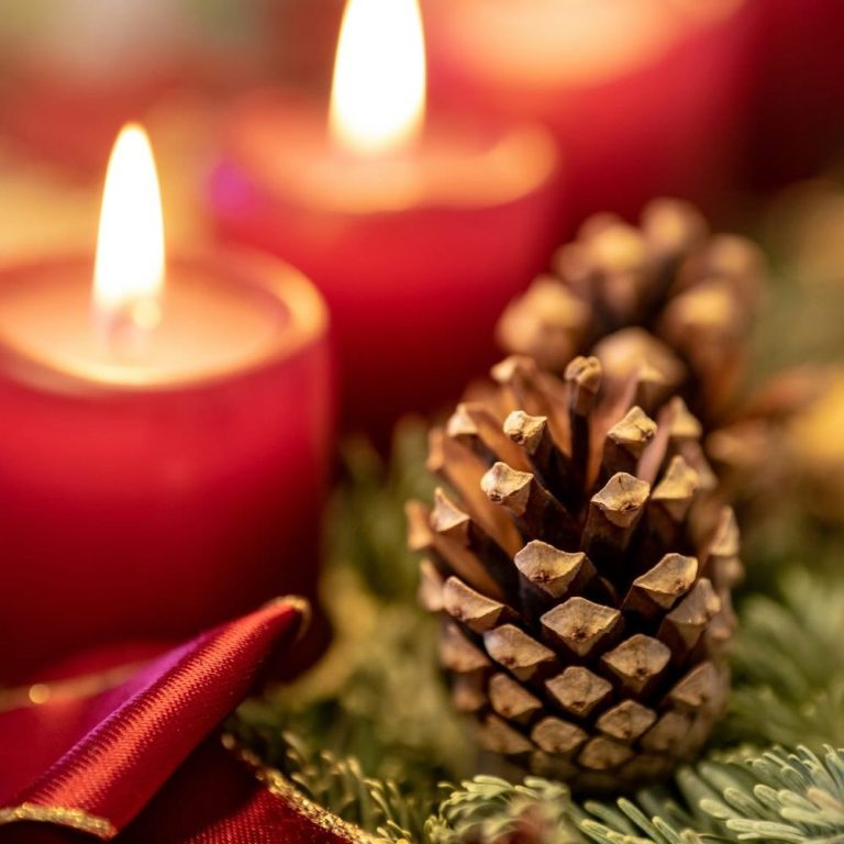Nahaufnahme eines Tannenzapfens vor brennenden roten Adventskerzen im Hintergrund.
