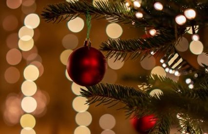 Nahaufnahme eines Tannenzweigs mit einer roten Christbaumkugel vor verschwommenen Lichtern – Symbol für einen achtsamen Advent und Ruhe.