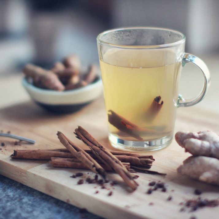 Holzbrett mit Ingwer, Kurkuma und Zimtstangen neben einem Glas Tee – wärmende Gewürze für die Ayurveda Vata-Checkliste.